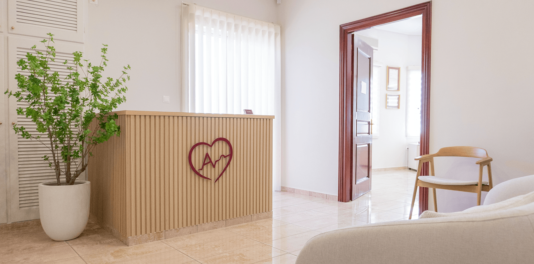 Χώρος υποδοχής ιατρείου με ξύλινο γραφείο, λευκή επιγραφή του ονόματος της γιατρού στο εμπρός μέρος και πράσινο φυτό δίπλα. Ο χώρος είναι φωτεινός και λιτός.